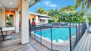 Une piscine avec une terrasse en bois, entourée d’une clôture, et des palmiers en arrière-plan.