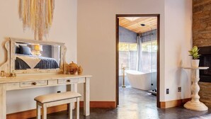 A room with a wooden desk, a bench, a mirror, a hanging decor, and a view of a bathroom with a bathtub. - The Soul of the River  (Three Rivers)