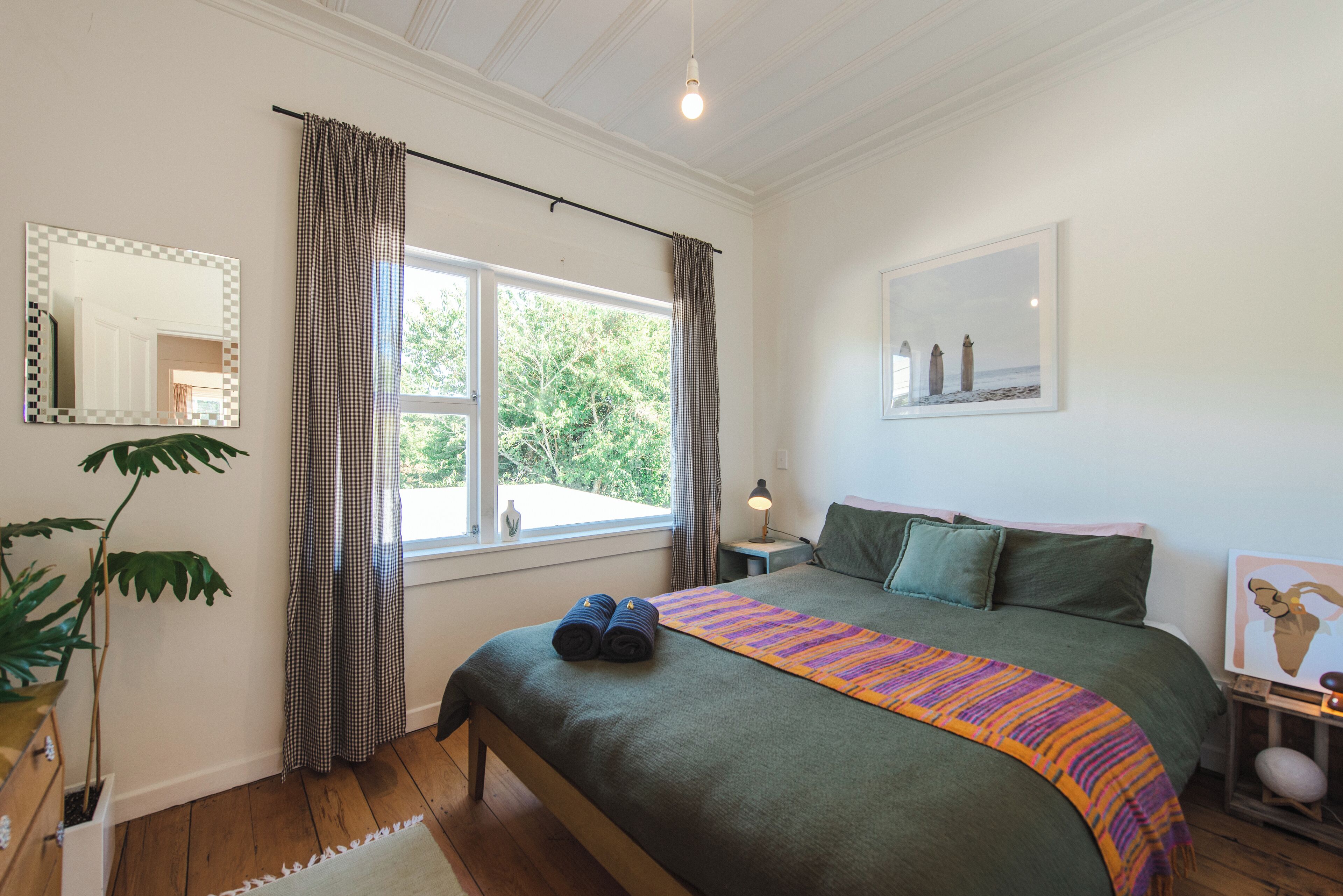 A bedroom with a large bed, a window with curtains, a bedside table with a lamp, and a framed picture on the wall.