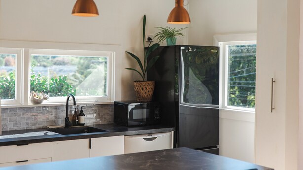 A modern kitchen with a black refrigerator, a microwave, and a sink. There are two pendant lights, a window with a view of greenery, and a potted plant on the counter.