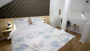 Een netjes opgemaakt bed met een bloemenprint deken, twee bedlampen en een klein rond tafeltje met een stoel.
