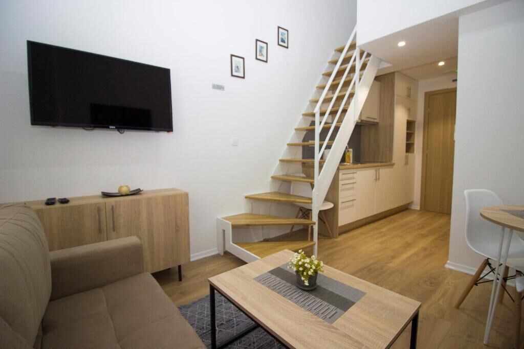 Un espace de vie moderne comprenant un canapé, une table basse avec un vase, un téléviseur fixé au mur et un escalier menant à une cuisine.