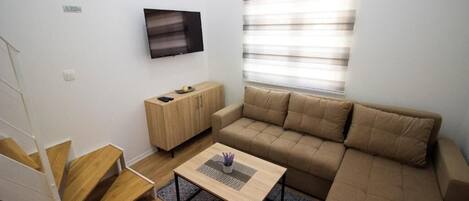 A modern living room with a sofa, a coffee table, a TV mounted on the wall, and a window with blinds.