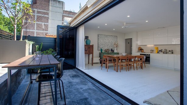 Une cuisine moderne avec des meubles blancs, un coin repas doté d’une table et de chaises en bois, et une porte vitrée donnant sur un patio extérieur avec une table et des chaises en bois.