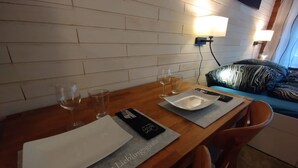Une table à manger en bois, avec un set de table, un menu noir et deux verres à vin. On aperçoit, à l'arrière-plan, un lit avec un oreiller à rayures zébrées.