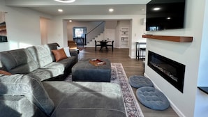 A modern living room with a sectional sofa, ottoman, fireplace, flat-screen TV, and a staircase leading to another level. - Spacious & Stocked: 2Kings+Hot Tub+Fire Pit+Family Fun (Albuquerque)