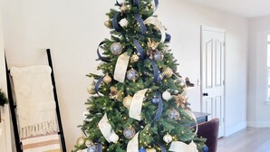 A Christmas tree decorated with ornaments and ribbons, placed in a living room with a wooden floor and a door in the background.