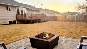A backyard with a fire pit, two lounge chairs, a wooden deck, and a house in the background.