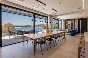 Un espace de restauration moderne avec une longue table et des chaises en bois, de grandes fenêtres offrant une vue sur la mer et les collines, et un mur en pierre agrémenté d’étagères.