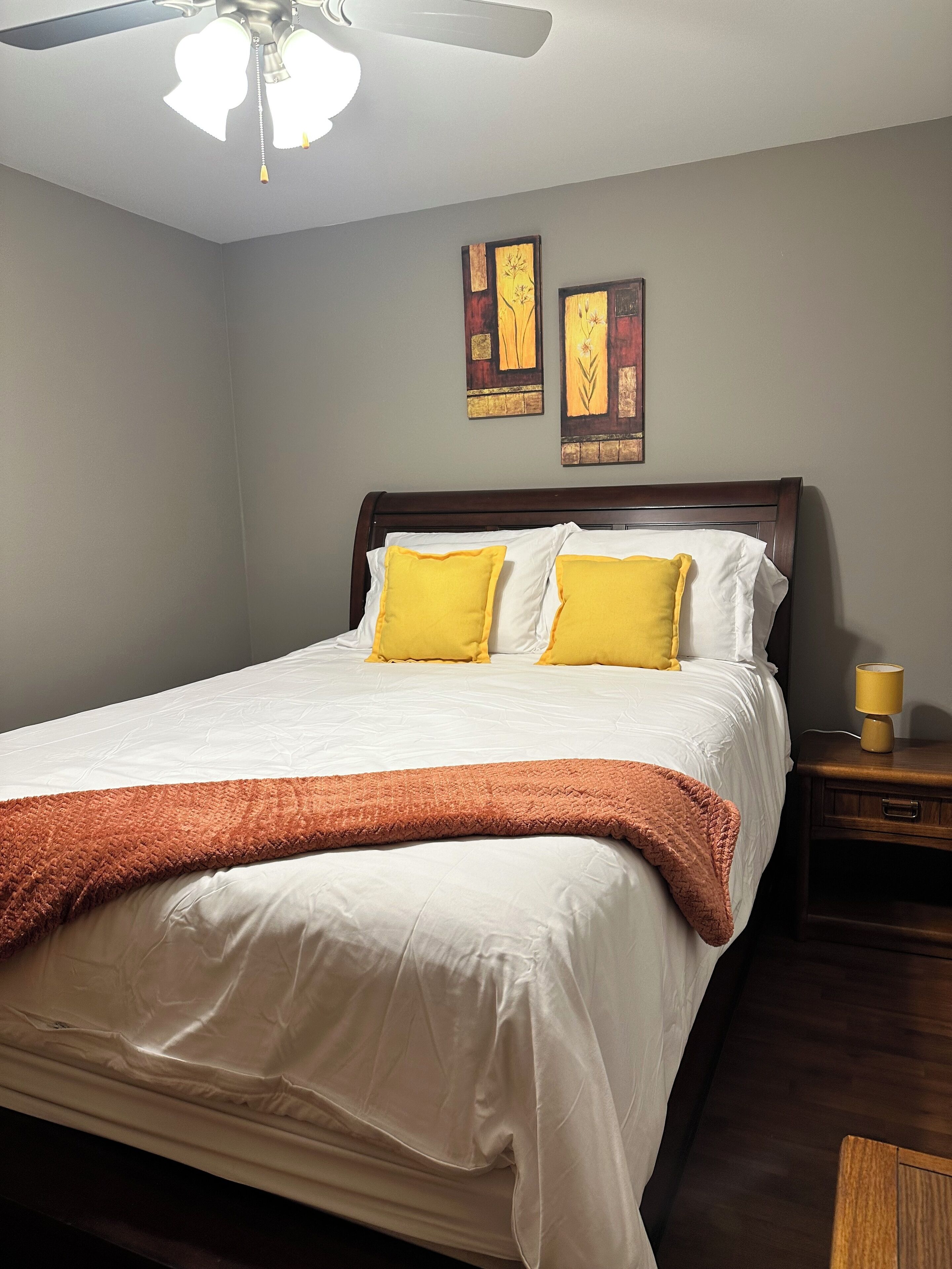 A neatly made bed with white and yellow pillows, a brown throw blanket, and a wooden headboard. There are two wall art pieces above the bed and a small bedside table with a lamp.