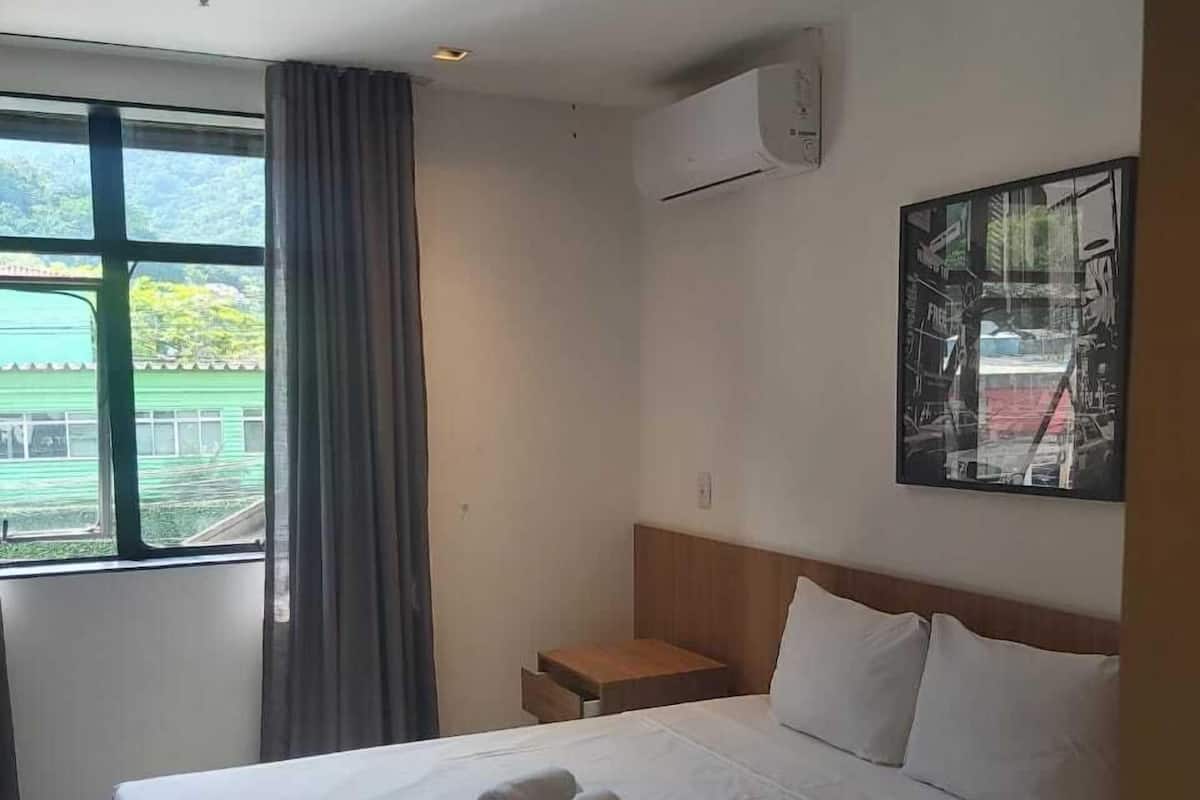 A hotel room with a large bed, a nightstand, a window with curtains, and a wall-mounted air conditioner.