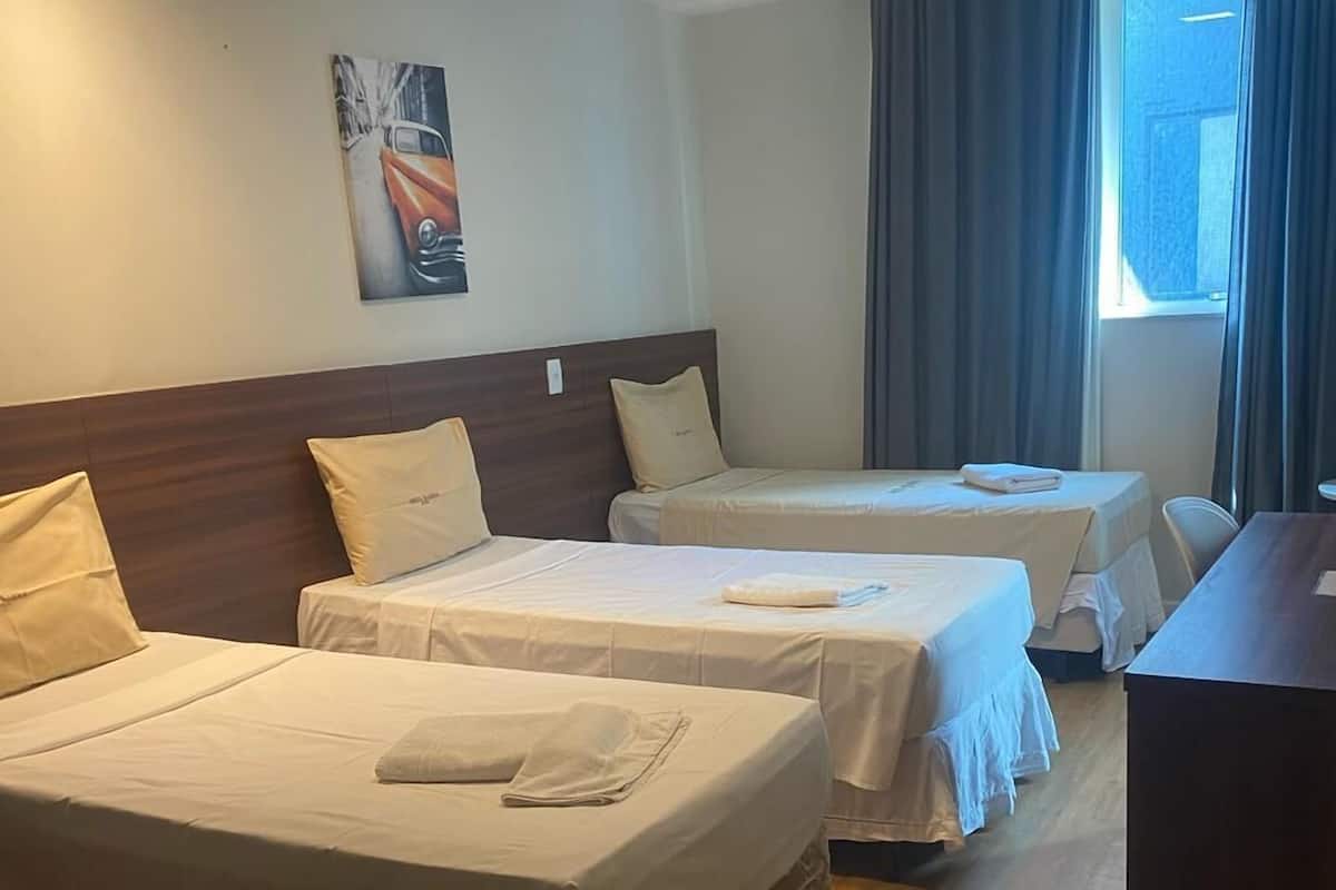 A hotel room with three beds, a wooden headboard, a painting on the wall, and a window with curtains.