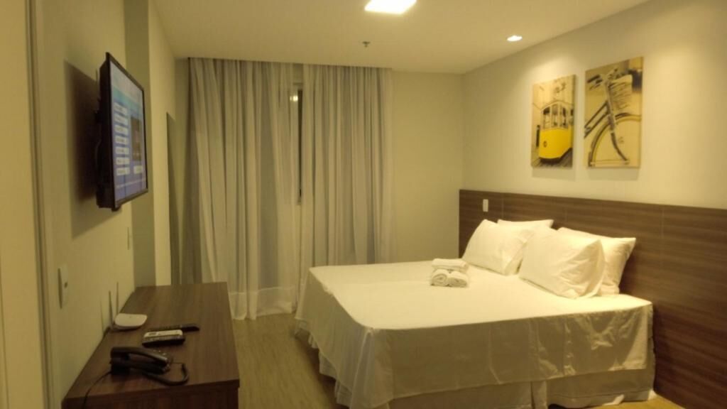 A hotel room with a bed, a desk with a phone and remote, a TV on the wall, and curtains.