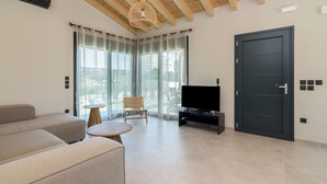 A modern living room with a sofa, a TV, a wooden ceiling, and large windows. - Elegant house with Private Pool (Schinias)