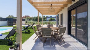 An outdoor patio with a round wooden table and chairs, a potted plant, and a view of a swimming pool and greenery. - Elegant house with Private Pool (Schinias)