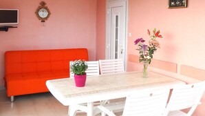 Une pièce avec un mur rose, une table blanche avec deux chaises, un canapé rouge, une plante en pot, une horloge murale et un tableau noir avec des dessins.