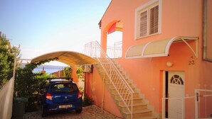 Une voiture bleue est garée dans une allée étroite qui mène à un bâtiment rose doté d’un escalier et d’une entrée couverte.