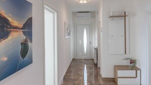 A hallway with a large painting of a boat on water, a door with a frosted glass panel, a radiator, and a small wooden side table with a plant. - Three Bedroom Apartment with Balcony and Sea View (Crikvenica)