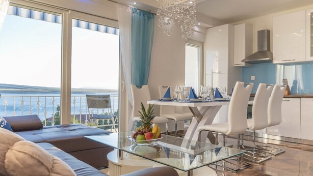 A modern living room with a glass coffee table, a sofa, and a dining area with white chairs and a blue tablecloth. - Three Bedroom Apartment with Balcony and Sea View (Crikvenica)