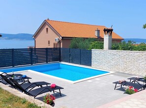 En swimmingpool med solstolar och en stenmur, ett hus med ett tak täckt av tegelpannor och utsikt över havet.