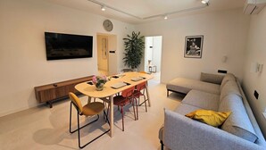 A modern living room with a sofa, a dining table with chairs, a TV mounted on the wall, and a large window. - Classic Two Bedroom Apartment with Balcony and Sea View (Crikvenica)