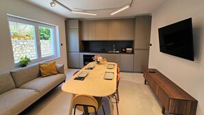 A modern kitchen with a built-in oven and stove, a dining table with chairs, a sofa, and a TV mounted on the wall. - Classic Two Bedroom Apartment with Balcony and Sea View (Crikvenica)