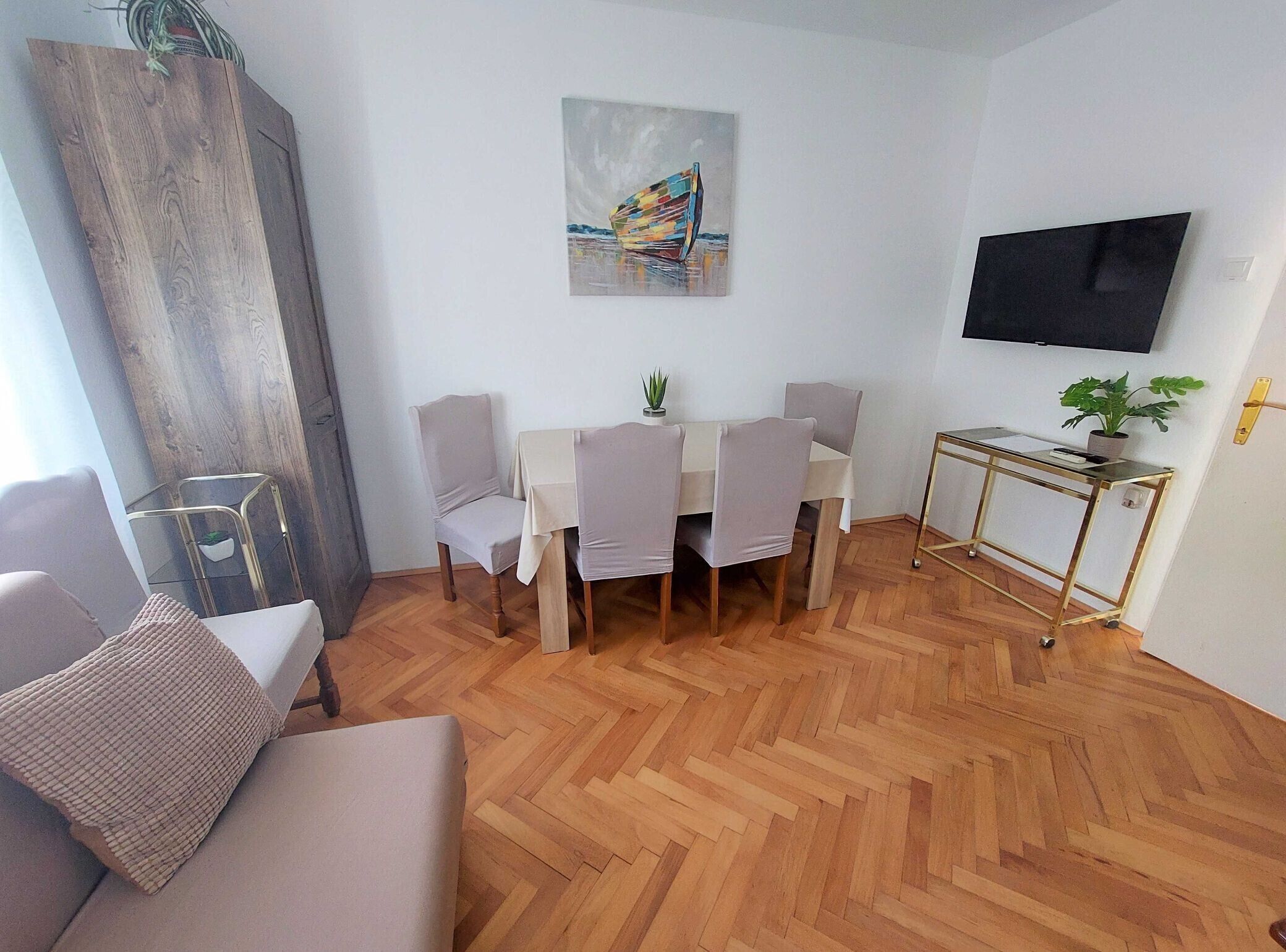 Una sala moderna con suelo de madera en espiga, una zona de comedor con una mesa blanca y cuatro sillas, un televisor de pantalla plana montado en la pared y una pequeña planta en maceta sobre una mesita de noche con marco dorado.