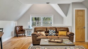 A loft-style living room with a brown leather sofa, a wooden coffee table, and a view of greenery through a large window.