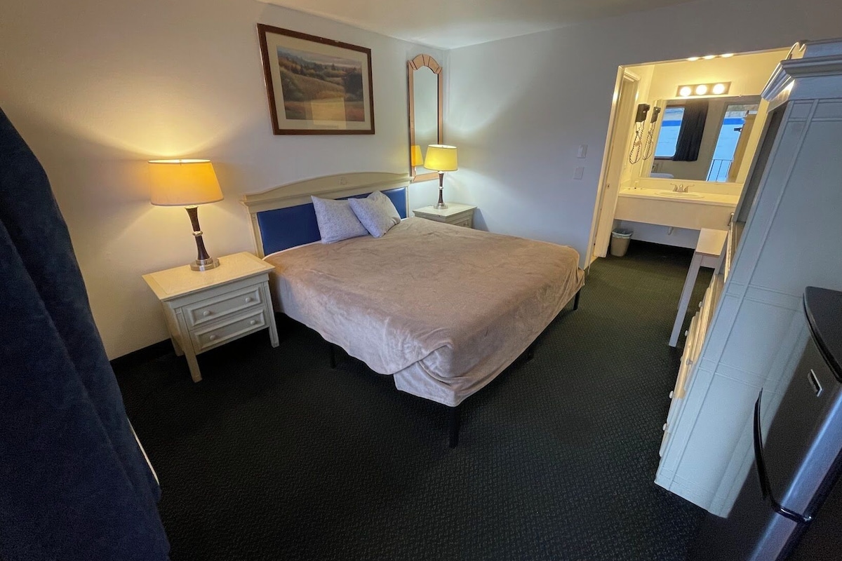 A hotel room with a bed, bedside lamps, a mirror, a small table, and a refrigerator.