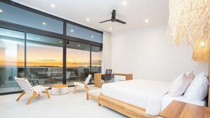 Ein modernes Schlafzimmer mit einem großen Bett, einem Schreibtisch, zwei Stühlen und Blick auf den Sonnenuntergang durch große Fenster.