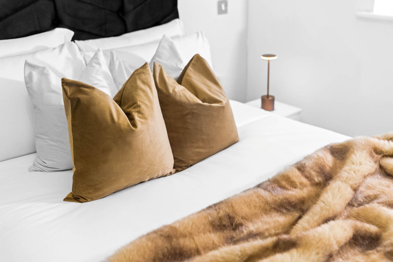 Un lit avec des oreillers blancs et bruns, un plaid en fausse fourrure et une lampe de chevet.