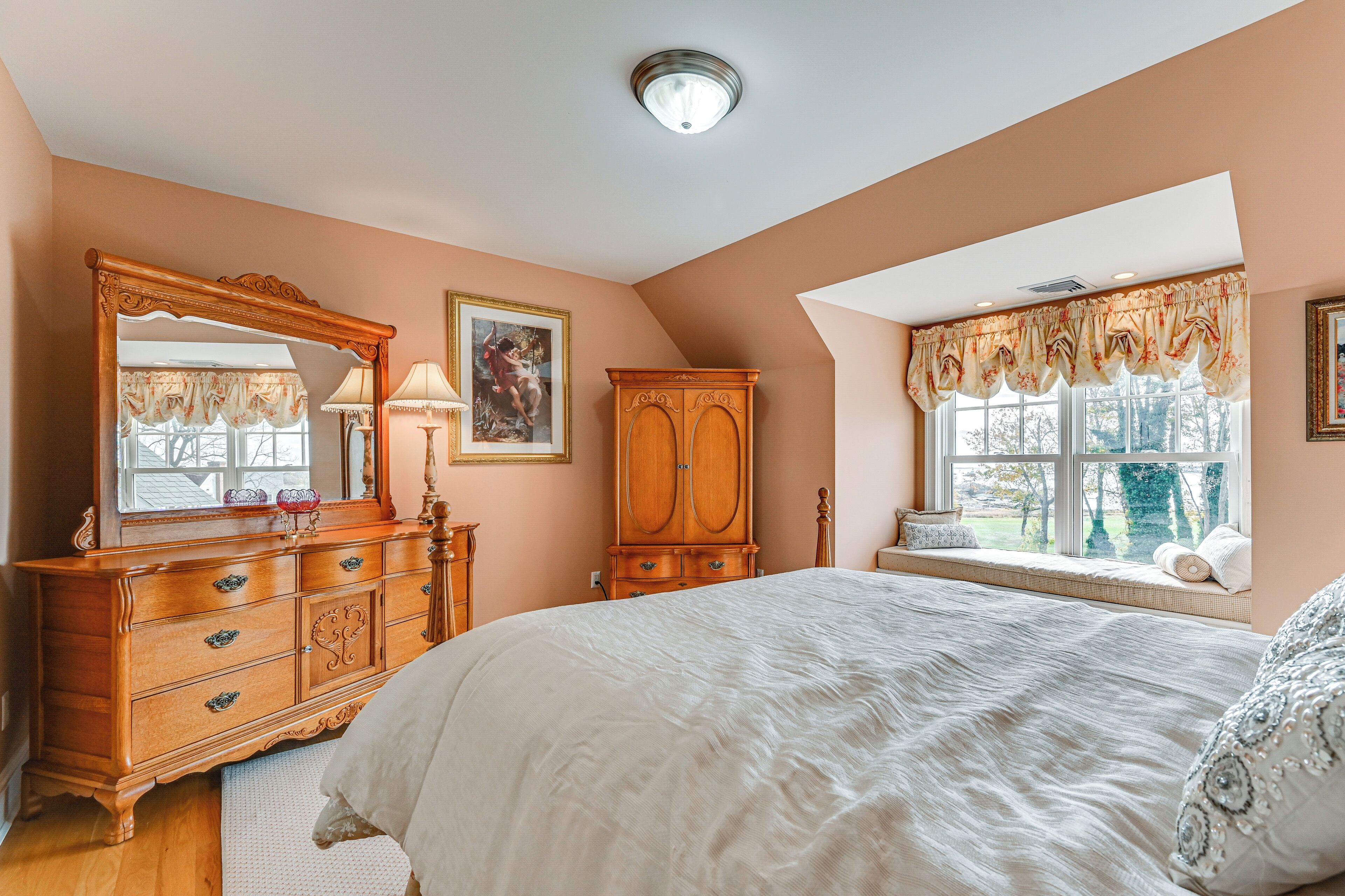 A bedroom with a large bed, wooden dresser, and a window with a view of trees.