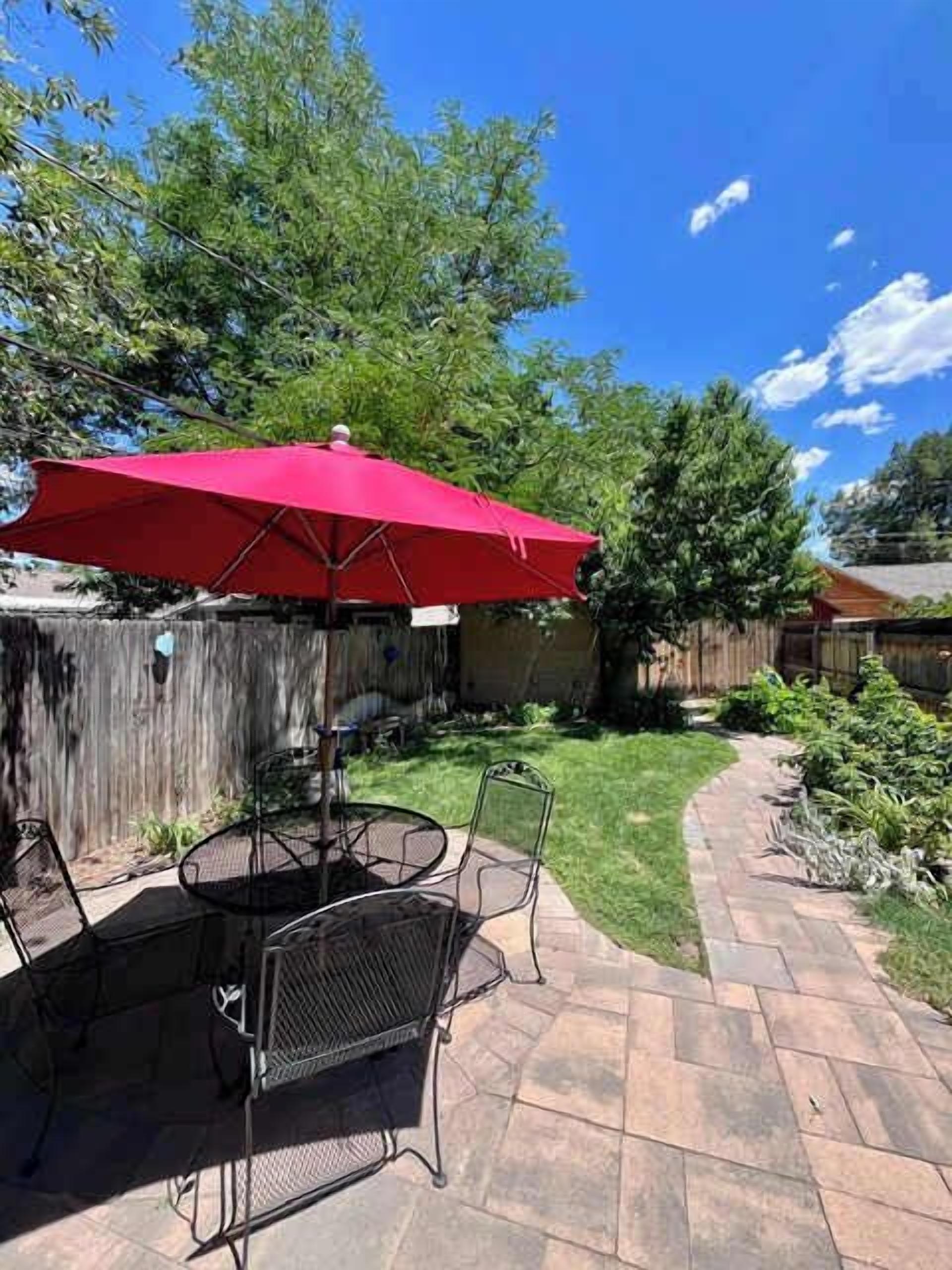Une terrasse extérieure avec un parasol rouge, des chaises en métal et un chemin piétonnier à motifs en chevrons.