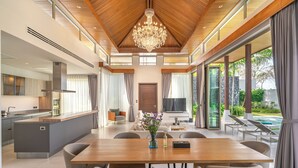Amplia sala de estar con techo de madera, candelabro, mesa de comedor con sillas y vista a una piscina y jardín.