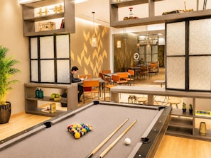 Une table de billard avec des billes, une personne utilisant un ordinateur portable et un coin repas avec des chaises orange.