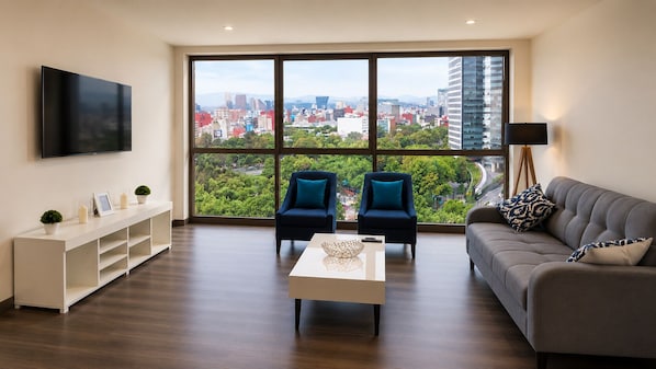 Un salon moderne avec une grande fenêtre, un canapé, deux fauteuils, une table basse et un téléviseur.