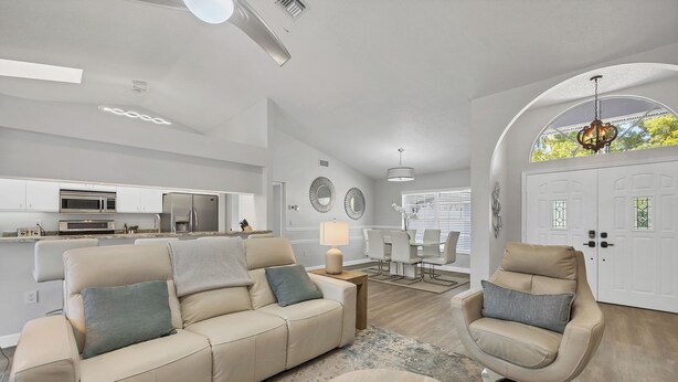 A modern living room with a beige sofa, a recliner, and a coffee table. There is a kitchen in the background with stainless steel appliances.