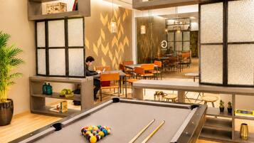 A pool table with balls, a person using a laptop, and a dining area with orange chairs.