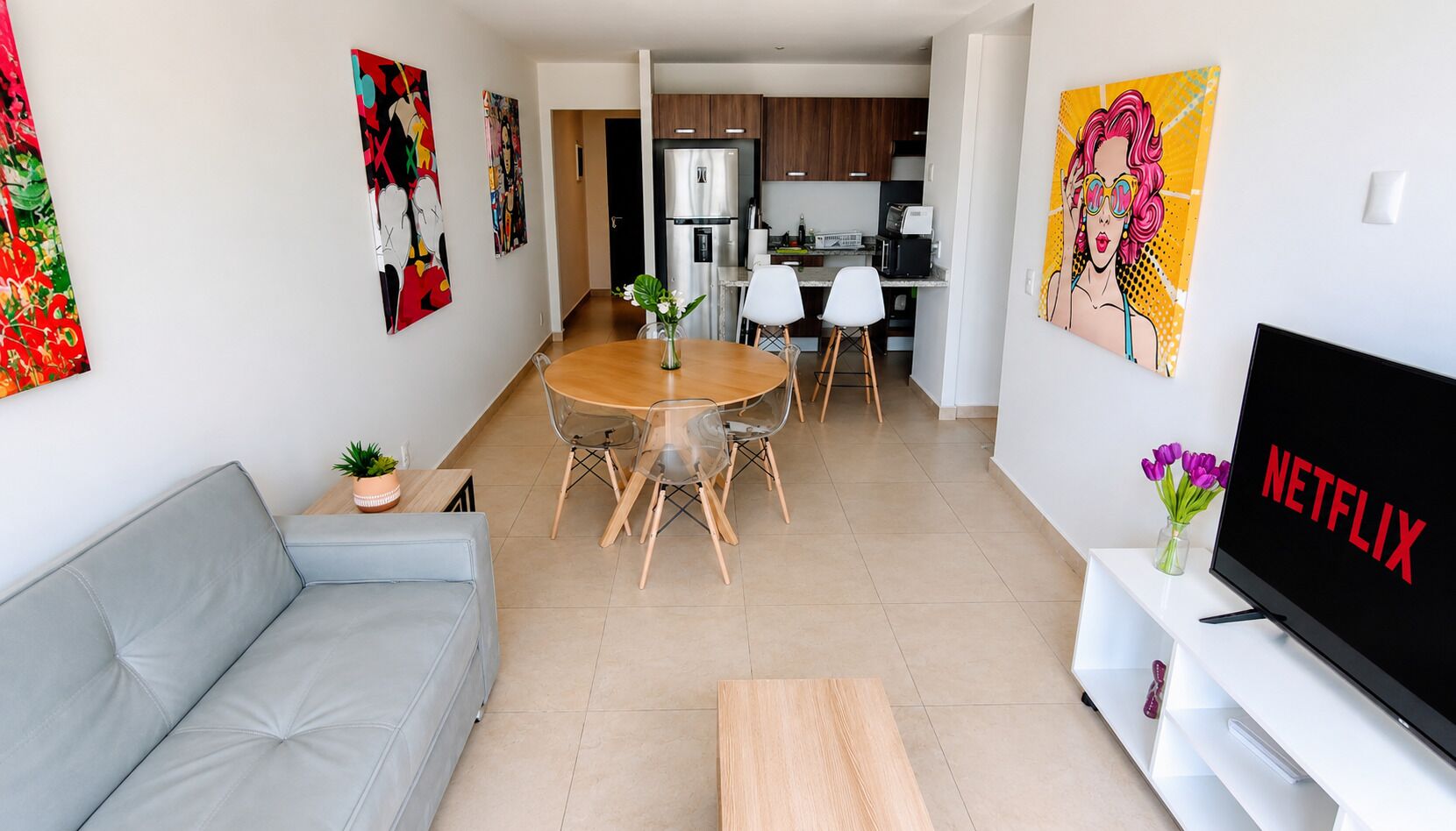 Una sala de estar moderna con un sofá gris, una mesa de centro de madera, un comedor con una mesa redonda y sillas, una cocina al fondo y un televisor mostrando el logotipo de Netflix.
