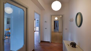 Un pasillo con suelo de baldosas, una mesita consola blanca con una figurita, un espejo redondo en la pared y una puerta que da a otra habitación.
