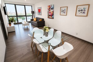 Una sala de estar moderna con una mesa de comedor de cristal, sillas blancas, un sofá gris y obras de arte en las paredes.