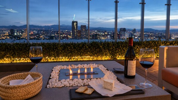 A rooftop terrace with a city view, a table with a cheese board, wine glasses, and a bottle of wine.