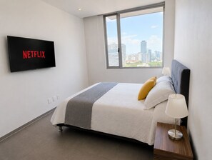 A hotel room with a bed, a TV mounted on the wall, a window with a city view, and a bedside table with a lamp.