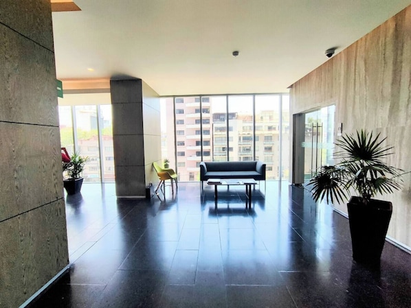 A spacious lobby with a dark tiled floor, a black sofa, a small table, and potted plants.