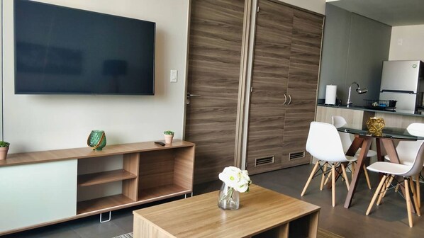 A modern living room with a flat-screen TV, wooden entertainment unit, a dining area with white chairs, and a kitchen with a refrigerator.