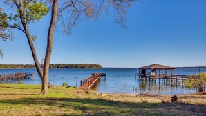 Escena junto al lago con un muelle de madera que se adentra en el agua, un pabellón cubierto y un gran árbol en primer plano.