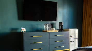 Un dormitorio con una pared azul oscuro, un televisor de pantalla plana montado sobre un tocador con tiradores dorados, un refrigerador antiguo y una cama con colcha blanca.