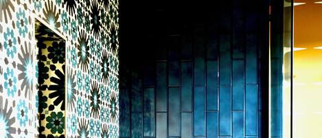 A bathroom with a patterned tile wall, a blue-tiled shower area, and a wooden bench.