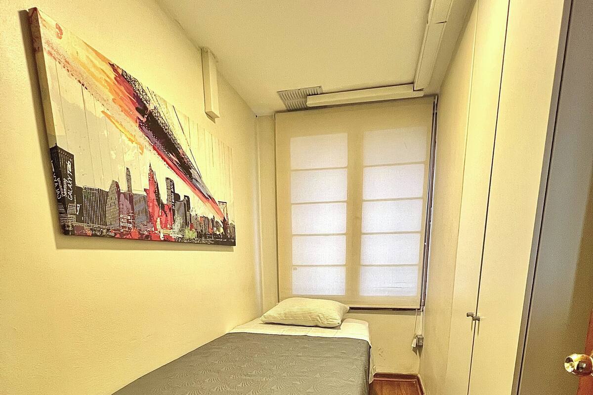 A small bedroom with a bed, a window with blinds, a door, and a cityscape painting on the wall.