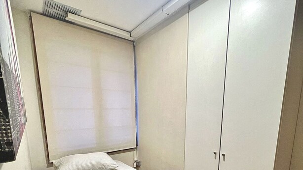 A small, single-bed room with a grey bedspread, a white pillow, a ceiling vent, and a wardrobe with two doors. - Hostel La Moneda (Santiago)